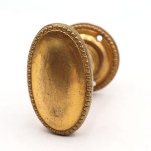 Op de afbeelding: Antieke messing deurknop met een ovale handgreep en een ronde achterplaat. De handgreep heeft een kralenrand en het messing heeft een warme, verouderde patina. De knop is ontworpen voor een deur.