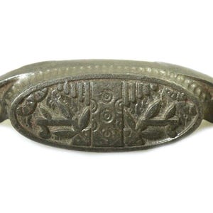 May include: A vintage cast iron drawer pull with an oval shape and a decorative floral design. The pull is a dark gray color and has two holes for mounting.