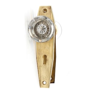 May include: A brass door knob with a clear glass crystal knob. The knob has a keyhole and is in the shape of a long rectangle.