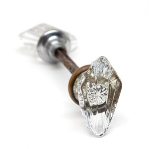 Pode incluir: Um puxador de porta de vidro transparente e facetado com formato de diamante. O puxador está preso a uma haste de metal enferrujado com um anel de latão. A outra extremidade da haste tem um puxador de vidro menor e retangular. O fundo é branco.