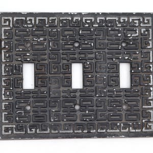 Könnte beinhalten: Eine rechteckige Dreifach-Lichtschalterabdeckung mit schwarzem Finish und abgenutzten silberfarbenen Kanten. Die Oberfläche ist mit einem sich wiederholenden geometrischen Mäander-Muster verziert und weist drei vertikale rechteckige Ausschnitte auf.