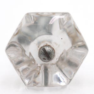 May include: Clear, faceted glass door knob with a hexagonal shape. The knob has a central screw for attachment and is designed with multiple facets to reflect light. A vintage hardware item.