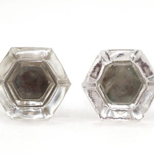 Puede incluir: Dos pomos de vidrio transparente, hexagonales, con aspecto esmerilado. Los pomos tienen forma de seis lados con un centro empotrado. El vidrio tiene una superficie ligeramente texturizada, sobre fondo blanco.