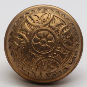 Op de afbeelding: Een gouden metalen knoop met een ingewikkeld bloemmotief. De knoop heeft een verhoogd midden met een kleiner bloemmotief en een omringende rand van bladeren en spiralen.