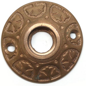 Puede incluir: Una placa metálica circular de color bronce, con un agujero central y dos agujeros más pequeños. La placa está decorada con un patrón repetitivo de diseños de hojas estilizadas alrededor del perímetro. El metal tiene un aspecto ligeramente envejecido.