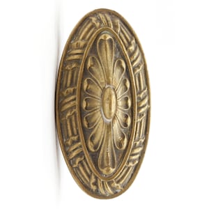 Puede incluir: Una perilla de puerta decorativa de forma ovalada de color dorado con un diseño floral en el centro y un patrón tejido alrededor del borde.