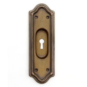 Könnte beinhalten: Ein antiker Schlüsselschlitzabdeckung aus Messing mit einem verzierten Design und einem Schlüsselschlitz in der Mitte.