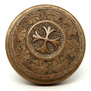 May include: A detailed, antique bronze door knob with an ornate floral and scroll design. The knob is round with a central medallion featuring a stylized floral motif. The bronze has a warm, aged patina.