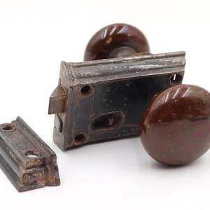 May include: A vintage door latch mechanism featuring two round, glossy dark brown ceramic knobs attached to a rectangular black metal lock box, accompanied by a separate matching black metal strike plate with two screw holes for mounting.