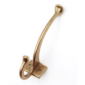 May include: A brass-toned metal wall hook featuring a dual-prong design with a long, curved upper arm and a shorter, lower hook. The base is a rectangular plate with two pre-drilled circular holes for mounting, finished in a brushed metallic texture.