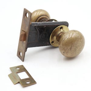 May include: Antique door hardware set featuring a brass doorknob, a black metal lock mechanism, and a separate strike plate. The doorknob has a rounded shape and a worn, aged appearance.