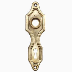 May include: A brass keyhole cover with an ornate design. The cover has two keyholes, one large and one small.
