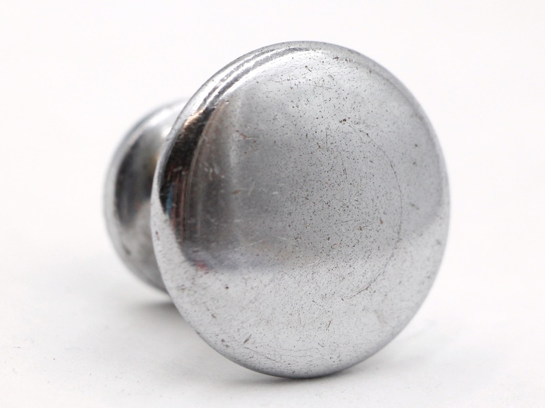 May include: A silver metal knob with a round, slightly textured surface. The knob has a small, raised lip around the edge.