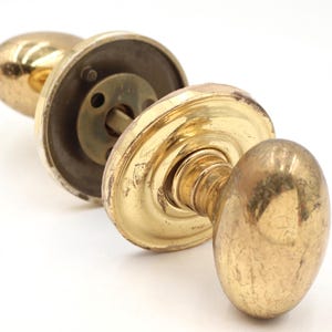 May include: A polished brass door knob set featuring two oval-shaped handles attached to circular mounting plates. The metal surface shows signs of wear and minor scratches, with a central spindle connecting the two sides of the hardware assembly.