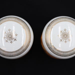 May include: Two white and gold decorative cabinet knobs. Each knob features a white center with a gold snowflake design and a gold border. The knobs have a round shape and are set against a black background.
