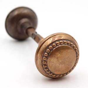 Könnte beinhalten: Antiker Türknauf aus Bronze mit rundem, strukturiertem Design. Der Knauf hat einen dekorativen Rand aus kleinen, erhabenen Punkten. Der Griff hat eine einfache, zylindrische Form, und das Gesamtdesign ist Vintage.