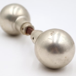 May include: A vintage door knob with a silver finish. The knob has a spherical shape on both ends, connected by a short, cylindrical piece. The connecting piece has a brown, possibly wooden, element. The background is white.