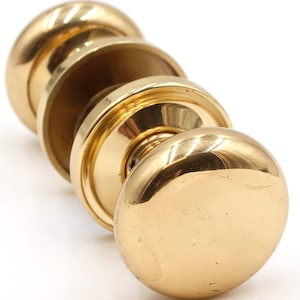 May include: Close-up of a set of antique brass door knobs. The knobs are round with a polished finish, reflecting light. The set includes a pair of knobs and a connecting piece, all in a warm, golden hue. Ideal for vintage home decor.