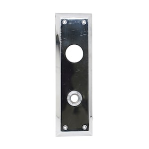 Pode incluir: Uma placa de porta retangular cromada com uma superfície polida. Possui uma grande abertura circular na parte superior e outra menor na inferior, com um anel decorativo. Quatro pequenos parafusos são visíveis, um em cada canto.