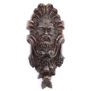 Può includere: Un decoro da parete in legno marrone scuro con un viso scolpito e dettagli decorativi. Il viso ha la barba ed è circondato da foglie e uva.