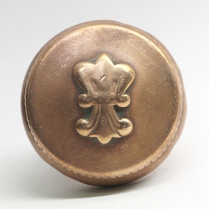 Puede incluir: Un tapón de botella redondo de color dorado con un diseño decorativo de flor de lis estilizado.