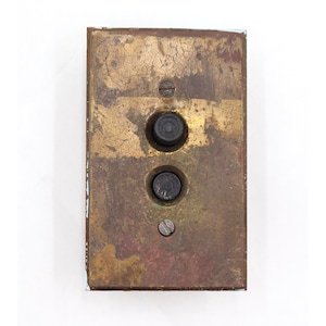 Może przedstawiać: Prostokątny, zabytkowy mosiężny przycisk dzwonka z dwoma czarnymi przyciskami i śrubą. Mosiądz ma zwietrzały, postarzany wygląd z przebarwieniami i patyną, co sugeruje wiek i użytkowanie.