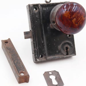 May include: An antique door lock set featuring a dark brown and red marbled glass doorknob. The set includes a black metal lock, a strike plate, and a separate metal piece, all showing signs of age and wear.