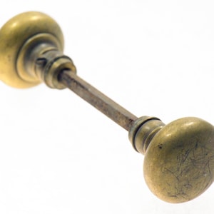May include: Antique brass door knob with an oval shape on each end, connected by a rectangular rod. The brass has a worn, aged appearance with visible scratches and discoloration, suggesting its vintage. The knob is set against a white background.