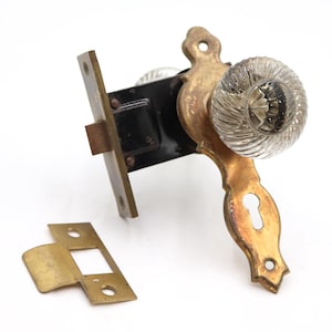 Könnte beinhalten: Ein Türgriff-Set mit einem klaren, geriffelten Glasknauf, der an einer verzierten, gealterten Messing-Rückenplatte mit Schlüsselloch befestigt ist. Der Mechanismus umfasst eine schwarze Metallfalle und ein separates, rechteckiges Messing-Schließblech.