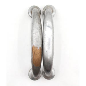 May include: Two vintage silver-toned metal door handles. Each handle has a curved grip and rounded ends. One handle shows exposed wood with a worn appearance, while the other is fully silver. The handles are set against a white background.