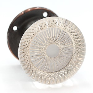 Op de afbeelding: Een heldere glazen deurknop met een decoratief, geometrisch patroon. De knop is bevestigd aan een ronde, donkerbruine metalen basis.