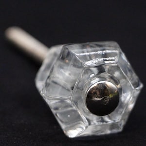 Puede incluir: Un pomo de cristal transparente con forma hexagonal y un centro de metal plateado. El pomo tiene un tornillo en la parte posterior para sujetarlo a una superficie.