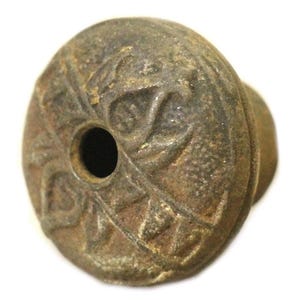 Może przedstawiać: Zbliżenie na mały, okrągły, metalowy przedmiot z otworem w środku. Przedmiot jest ozdobiony wzorem kwiatowym i ma szorstką, fakturowaną powierzchnię.