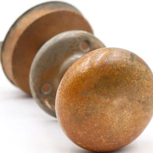 May include: Three antique door knobs in varying sizes and shapes, arranged in a diagonal line. The knobs have a weathered, aged appearance with a mix of brown and copper tones, suggesting they are made of metal. The background is plain white.