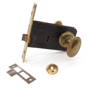 May include: Antique brass door lock set with a round knob, a square strike plate, and a separate keyhole cover. The lock has a dark brown finish and a keyhole on the front.