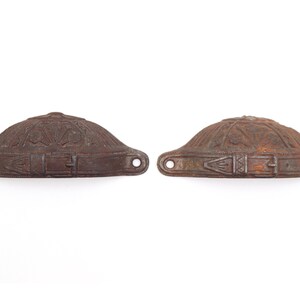 May include: Two antique, bronze-coloured metal drawer pulls. Each pull has a domed, hat-like shape with ornate floral and geometric designs. The pulls feature a buckle detail and two mounting holes.