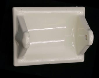 May include: White ceramic toilet paper holder with a simple design. The holder has a rectangular base and a curved arm to hold the roll.