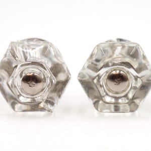 Puede incluir: Dos pomos de cristal transparente con un diseño hexagonal facetado. Cada pomo presenta un centro de metal plateado. Los pomos se muestran sobre un fondo blanco, resaltando su transparencia y forma.