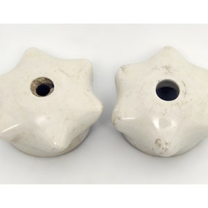 Puede incluir: Dos pomos de armario en forma de estrella, de color blanquecino. Cada pomo tiene seis puntas y un agujero central. Los pomos tienen un aspecto ligeramente envejecido, con sutiles variaciones de color y textura. Probablemente sean de cerámica.