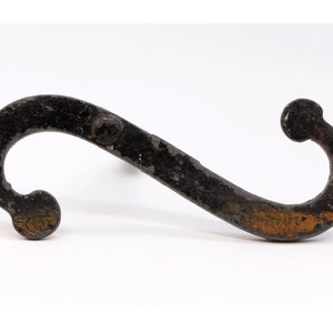 May include: A rustic, black metal hook with an S-shaped design. The hook has a textured surface with visible wear and tear, and a small, rounded knob at each end. The metal has areas of orange-brown discoloration.