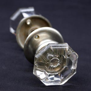 May include: A pair of vintage door knobs with clear, faceted glass knobs and brushed metal backplates. The knobs have an octagonal shape and are set against a dark background, showcasing their reflective qualities. A classic hardware item.