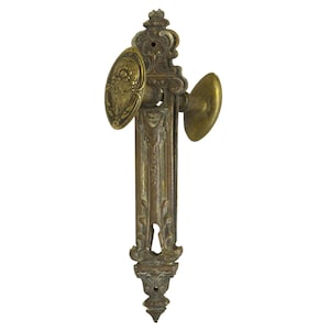 Op de afbeelding: Een antieke messing deurknop met ingewikkelde bloemmotieven en een sleutelgat. De knop heeft een ronde knop aan de rechterkant en een ovale knop aan de linkerkant.