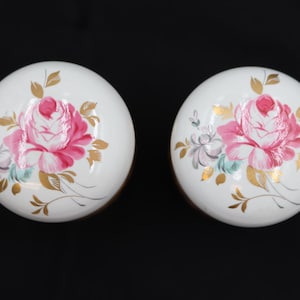 May include: Two white, round decorative knobs with hand-painted floral designs. Each knob features a large pink rose with gold and green accents. The knobs are set against a black background.