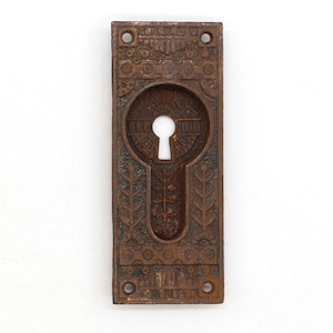 Może przedstawiać: Antyczna, ozdobna osłona otworu na klucz wykonana z żeliwa. Osłona ma kształt prostokąta z otworem na klucz w środku. Osłona jest ciemnobrązowa.