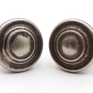 Peut inclure: Deux boutons de meuble argentés antiques avec un design circulaire et superposé. Les boutons présentent une finition vieillie, suggérant l'âge et l'utilisation. Le matériel est posé sur un fond blanc, mettant en valeur les détails.
