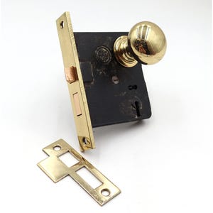 May include: A vintage door lock set with a brass finish. The set includes a black metal lock body, a brass door knob, a brass strike plate, and a brass faceplate. The lock is designed for a door and is likely from an older home.