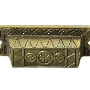 Może przedstawiać: Mosiężny uchwyt do szuflady z geometrycznym wzorem. Uchwyt ma prostokątny kształt z podniesioną częścią środkową, na której znajduje się stylizowany motyw kwiatowy. Krawędzie uchwytu są ozdobione powtarzającym się wzorem trójkątów i rombów.