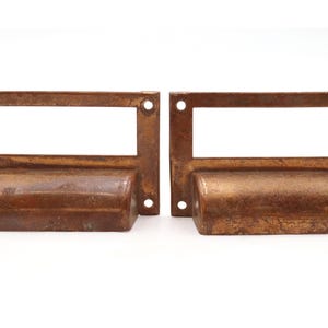 May include: Two rectangular drawer pulls made of aged, weathered bronze metal. Each pull features a central horizontal handle grip and a rectangular label holder frame above it, with four circular screw holes positioned at the corners.