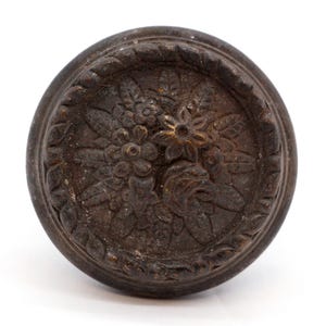 May include: A dark brown, round, decorative knob with a floral design. The knob has a raised, textured border.
