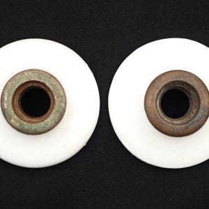 May include: Two circular white ceramic insulators, each featuring a central threaded metal insert. The metal components exhibit a weathered, oxidised brown and greenish patina, contrasting with the smooth, glossy white finish of the ceramic discs.